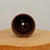 A top-down view looking into a circular, dark brown unglazed ceramic bonsai pot. The interior of the pot is smooth, featuring a prominent central drainage hole and four smaller peripheral holes, which are crucial for ensuring healthy drainage and air circulation for bonsai roots. The pot rests on a thick, natural wood cross-section with a visible light-toned grain. The background is a solid, neutral off-white, providing a clean contrast that emphasizes the rich, earthy color and functional design of the ceramic pot.