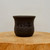 A studio-shot, eye-level close-up of a small, dark brown, unglazed ceramic bonsai pot. The pot features a classic design with a flared rim and a rounded body that tapers slightly toward the base. Its matte surface is adorned with intricate black Chinese calligraphy characters of varying sizes, which are etched into the side of the pot. A set of larger, bold characters dominates the center, while smaller, more delicate inscriptions and a traditional-style signature seal are positioned below and to the side. The pot sits on a thick, circular piece of light-toned natural wood, showcasing its organic grain and texture. The entire composition is set against a clean, neutral, off-white background, emphasizing the pot's earthy tones and artistic, etched details.