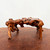 An intricately detailed, low-profile bonsai root stand sits centered on a weathered wooden surface against a plain, off-white background. Crafted from polished natural wood, possibly a root or burl, the stand features a rich palette of warm amber and golden-brown tones, punctuated by dark chocolate and blackish knots and grain patterns. Its form is organic and gnarled, with a highly textured surface that has been polished to a glossy sheen. The stand is supported by four uneven, trunk-like legs that emerge from its body in an asymmetrical fashion, giving it the appearance of a miniature, weathered bridge. The top surface, while relatively level for display purposes, follows the natural, irregular contours of the wood. This decorative base is designed to provide an elevated and aesthetically pleasing platform for displaying bonsai trees, suiseki (viewing stones), or other fine art objects, blending natural rustic charm with a refined, traditional finish.