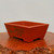 A rectangular, unglazed reddish-brown bonsai pot sits on a polished wooden slab with a natural, rustic edge. The planter has a tapered design, wider at the top than the bottom, and is supported by four small, integrated feet. On its front-facing side, there is a detailed, dark-toned etching of a serene mountain landscape featuring stylized trees and rolling hills. To the left of the landscape scene, small calligraphic characters are delicately engraved into the clay. The pot's matte, earthy finish, characteristic of high-quality unglazed ceramic or terracotta, contrasts with the glossy surface of the wooden stand. The entire piece is set against a plain, light-colored wall, drawing focus to the craftsmanship and traditional aesthetic of this etched bonsai container.