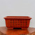 A rectangular, unglazed reddish-brown bonsai pot sits on a polished, dark-grained wooden slab, which is placed on a rustic, live-edge wood surface. The pot features a slightly flared rim and is supported by four small, block-style feet. On its front face, an extensive and intricate series of black Chinese calligraphy characters is etched, arranged in seven vertical columns that cover most of the surface. A smaller vertical signature and seal are visible on the far left side of the inscription. The entire scene is set against a plain, light grey background, highlighting the earthy texture of the clay and the fine detail of the etched poem. This type of etched Yixing-style container is a classic choice for traditional bonsai presentation.