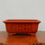 A detailed photograph of an unglazed, rectangular bonsai pot made from a warm, reddish-brown clay, characteristic of traditional Yixing or Zisha pottery. The pot features a subtly flared rim and four small, sturdy feet at its base. On its front surface, several vertical columns of elegant, black etched Chinese calligraphy add a classic artistic touch. The pot is positioned centrally on a dark, polished wood grain surface, set against a clean and minimalist off-white background that emphasizes its shape and decorative details.
