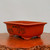 A high-angle studio shot features a rectangular, unglazed bonsai pot made from warm, reddish-orange clay. The pot has a wide, slightly flared rim and sits on four small, sturdy feet. Its front side is decorated with a traditional landscape scene etched in black ink, depicting layered mountains, stylized pine trees, a small boat on a body of water, and a traditional hut or building. A few small calligraphic characters are etched in the upper left corner of the scene. The pot is positioned on a polished, dark-toned wood surface with a rough, natural edge, against a plain, light-gray background. This unglazed ceramic planter is a classic example of an etched bonsai pot, combining functional design with artistic detail.