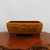 A rectangular, unglazed ceramic bonsai pot sits centered on a polished, dark brown wooden slab with a natural, rough-hewn edge. The planter features a two-tone color scheme: the main body is a warm golden-yellow or mustard hue, while the interior, the rim, and the blocky feet are a contrasting reddish-brown terracotta color.

The front face of the pot is decorated with delicate black ink etchings depicting a traditional mountain landscape. On the upper left side, there are several Chinese characters. Below the calligraphy, a small hill with vertical fence-like lines is sketched. To the right, a larger scene unfolds, showing layered mountain peaks, rolling hills, clusters of tall trees, and a small group of buildings nestled in the valley. The entire scene is set against a plain, light-gray background, highlighting the intricate detail of the etched artwork and the clean lines of the ceramic planter.