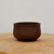 A side-view studio photograph of a small, round unglazed ceramic bonsai pot in a deep, dark brown chocolate tone. The planter features a smooth, matte finish with slightly bulbous, rounded sides and a sturdy, recessed base. It is centered on a thick, light-toned natural wood cross-section slab, set against a clean, neutral off-white background. This minimalist ceramic container is designed for bonsai cultivation, offering a classic and understated aesthetic for potting small trees or succulents.