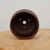 An interior, top-down view of a deep, round, unglazed ceramic bonsai pot in a rich, dark chocolate-brown color. The pot's smooth, matte finish is showcased as the camera looks directly into its deep basin, revealing several holes at the bottom essential for plant health: one large central drainage hole and three smaller holes positioned around it for securing bonsai wire. The pot is centered on a light-colored, natural wood surface, set against a plain, neutral off-white background. This type of sturdy, unglazed planter is designed to provide the necessary aeration and drainage required for healthy bonsai tree root systems.
