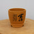 A detailed view of an unglazed, tan ceramic bonsai pot featuring elegant black Chinese calligraphy etched on its side. The pot has a classic tapered cylindrical shape with a smooth, matte finish and a slightly flared rim. Its decorative footed base includes small scalloped cutouts for both aesthetic appeal and drainage. 

On the side of the pot, bold calligraphy characters for "舍得" (Shědé) are painted in fluid black ink, signifying the philosophy of "giving and receiving." Smaller vertical columns of script and a tiny red seal mark accompany the main text, adding to its traditional Yixing-style aesthetic. The pot is positioned on a natural, light-colored wooden surface against a neutral, light-gray background, emphasizing its earthy texture and artisanal craftsmanship.