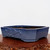 A low-profile, rectangular Yixing bonsai pot with a rich blue glaze, featuring elegantly notched corners and a shallow depth. The ceramic planter is finished in a vibrant indigo or sapphire blue with subtle tonal variations that give the surface a handcrafted, textured appearance. On the front-facing panel, there is a raised, embossed floral or pine needle motif. The pot sits on four low, integrated feet for stability and drainage. Its interior is a dark, unglazed earthy brown, characteristic of traditional Yixing clay. The planter is displayed on a polished, warm-toned wooden slab with a natural, textured burl edge, set against a clean, off-white background.