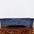 A low-profile, rectangular Yixing bonsai pot featuring a deep cobalt blue glaze with a subtle, matte finish. The front panel of the ceramic planter is decorated with a raised relief carving of two stylized, clustered flowers attached to a winding branch. The pot has a clean, geometric silhouette with indented corners and sits on small, sturdy feet at each corner. It is photographed on a polished, reddish-brown wooden display stand with a rustic, natural edge against a stark white background, highlighting the rich color and artisanal detail of the glazed pottery.
