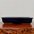 A side-profile photograph of a rectangular Japanese bonsai pot featuring a deep, glossy navy blue glaze. The shallow and wide planter, typical of traditional bonsai styles, sits elegantly on a polished, reddish-brown wood surface with a textured, natural edge. The smooth ceramic finish of the pot reflects soft light, highlighting its clean lines and subtle outer lip at the top. The pot is elevated slightly by a recessed base, designed to facilitate proper drainage for a living tree. Set against a minimalist off-white background, this high-quality blue bonsai pot is presented as a sophisticated and durable choice for any bonsai enthusiast looking for a classic container for their miniature trees.
