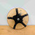 A detailed underside view of a professional bonsai turntable workstand resting on a wooden workbench. The top is a circular, light-colored wooden platform with a clean finish. Centered on the underside is a sturdy, five-armed star-shaped base in matte black, which provides a wide and stable foundation. Each arm of the base is fitted with a square black rubber foot to prevent slipping and protect surfaces. A central swivel mechanism, consisting of a square metal plate, is sandwiched between the wooden top and the star base, allowing for smooth, 360-degree rotation. A silver metal eyelet screw is visible near the center, functioning as a locking pin to secure the turntable in a desired position. This heavy-duty tool is essential for bonsai enthusiasts, enabling them to easily rotate their trees for precise pruning, wiring, and styling from every angle.
