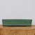 A side view of a ten-inch rectangular Yixing bonsai pot with a soft, matte celadon green glaze. The low-profile planter features slightly rounded corners and horizontal ridge detailing near its base, which rests on four small, shallow feet. The top rim has a dark brown, unglazed edge, revealing the natural color of the Yixing clay underneath. The pot is centered on a thick, light-toned wooden cross-section, highlighting its handcrafted ceramic texture against a clean, neutral white background.