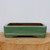 A 10-inch rectangular Yixing bonsai pot featuring a smooth, glossy sage green glaze. The planter has a traditional aesthetic with rounded corners and a decorative double-molded lip around the lower edge. It sits on four small, integrated feet for stability and drainage. The pot is displayed on a rustic, cross-cut wooden slab against a neutral, light-colored background. The interior of the pot shows the natural, unglazed brown Yixing clay, emphasizing its authentic construction and quality for bonsai cultivation.