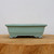 A side-view photograph of a seven-inch rectangular Yixing bonsai planter with a pale mint green or celadon glazed finish. The pot features a wide, flat rim and a slightly tapered body that sits atop four decorative, scroll-carved feet. A subtle brownish-red line accents the edges of the rim and the ornate details on the feet, giving it a traditional feel. The planter is centered on a thick, natural wood slab with a visible grain, set against a plain, off-white background. This elegant glazed bonsai pot is a classic choice for showcasing small to medium-sized trees.