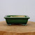 A side-view photo of a rectangular, seven-inch Yixing bonsai pot with a rich, glossy forest green glaze. The planter features a slightly flared rim and is supported by four decorative, scroll-shaped feet that show hints of darker glaze pooling in their crevices. The interior of the pot remains unglazed, revealing the natural, pale-beige color of the Yixing clay. The pot is positioned in the center of a thick, rustic wood slice that acts as a pedestal, all set against a clean and neutral off-white background. The soft, even lighting highlights the smooth, reflective finish of the glazed ceramic, emphasizing its elegant form.