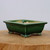 A detailed product shot of a seven-inch rectangular Yixing bonsai pot, showcasing its vibrant emerald green glazed finish. The ceramic planter features a slightly flared rim and is supported by four ornate, decorative cloud-style feet that reveal glimpses of the natural tan clay underneath the glossy glaze. The interior of the pot remains unglazed in a neutral tan color, while the exterior has a smooth, reflective surface. It is displayed on a light-colored wooden block against a minimalist, pale gray background, emphasizing the elegant craftsmanship and rich color of the glazed bonsai pot.