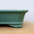 A detailed profile shot showcasing the side and corner of a teal-colored Yixing bonsai planter. The pot features a glossy, smooth glaze with a dense brown speckled pattern, creating a textured appearance similar to a robin's egg finish. The design includes a gracefully flared upper lip and a sturdy, decorative foot with rounded edges. The ceramic pot rests on a light tan surface against a plain white background, highlighting its color and craftsmanship.