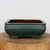 A side-on view of a nine-inch Yixing bonsai planter featuring a rich teal-green glaze with a subtle mottled texture. The ceramic pot has a rectangular form with elegant rounded corners and slightly flared sides. A thin horizontal ridge circles the upper portion of the planter, adding a touch of classical detail. It rests on four low, integrated rectangular feet. The glazed exterior is glossy, while the interior shows the natural, unglazed brownish clay. The pot is showcased on a rustic wooden slab, which sits against a clean, off-white background, highlighting the planter's vibrant color and clean lines.