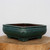 An artisanal nine-inch Yixing bonsai planter is featured in a close-up, showcasing its rich teal-green glaze. The rectangular ceramic pot has softly rounded corners, slightly bowed sides, and a prominent top rim. Its glaze is deep and lustrous, with subtle variations and light, speckled "frosted" textures near the upper edges that highlight its handmade character. 

The planter is elevated by four sturdy, blocky feet at each corner. It is positioned on a rustic, light-colored wooden slab with visible natural grain and bark texture along the edge. The entire scene is set against a clean, minimalist off-white background, emphasizing the elegant form and vibrant color of the glazed bonsai pot.