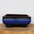 An image of a nine-inch rectangular glazed Yixing bonsai pot featuring a deep cobalt blue finish with a dark, almost black, gradient around the upper rim and feet. The planter has a sleek, low-profile silhouette with rounded corners and sits atop a rustic wooden slice against a neutral white background. Its vibrant blue glaze provides a sophisticated contrast, making it an ideal choice for showcasing the lush green foliage of a bonsai tree.