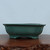 A profile view of a shallow, rectangular teal-glazed Yixing bonsai pot resting on a polished dark brown wooden display stand. The ceramic container features a sophisticated lobed or scalloped design along its sides, known as a mokkou shape, finished in a matte turquoise glaze with subtle color variations. A thick, outward-flaring rim defines the top edge, while the pot is supported by four integrated cloud-style feet that elevate it above the surface. The scene is set against a clean, light-blue background, emphasizing the artisan craftsmanship and elegant proportions of the Yixing pottery.