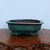 A rectangular Yixing-style glazed bonsai pot in a muted teal-green color sits on a polished dark wood stand. The pot's exterior features a soft, matte finish with subtle variations in the glaze, giving it an aged, ceramic appearance. It has an elegant shape with rounded corners, gently fluted sides, and a flat, outward-extending rim. Four small, integrated feet support the pot. The dark, unglazed clay interior is visible, indicating its suitability for bonsai plant health and drainage. The pot is centered on a glossy brown wooden slab with a natural, rough-hewn edge, all set against a neutral, light-blue background that emphasizes the pot's serene color and traditional craftsmanship.