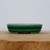 A side profile photograph of a shallow, oval-shaped Yixing bonsai pot finished in a vibrant emerald green glaze. The ceramic planter features a subtle, fine crackle pattern on its surface and a distinctive darker green horizontal groove encircling its midsection. This glazed bonsai pot has a slightly rounded rim and is elevated by small, integrated feet designed to allow for proper drainage. The pot sits on a rustic, light-brown wooden slab with visible grain, contrasted against a minimalist, off-white background that emphasizes its elegant form and rich color.
