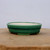 A shallow, oval-shaped Yixing glazed bonsai pot rests on a light-colored wooden surface against a plain white background. The ceramic planter features a vibrant, multi-toned emerald green glaze with darker green detailing along its stepped, double-rimmed exterior. The interior of the pot is a contrasting pale cream color. It is supported by small, integrated feet that lift it slightly off the surface. This classic green glazed bonsai pot is designed for small to medium-sized bonsai trees, blending traditional craftsmanship with a clean, professional aesthetic.