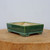 A rectangular Yixing glazed bonsai pot in a deep forest green color is showcased in a close-up, eye-level shot. The ceramic planter features a glossy finish that catches the light, with a slightly flared rim and a matching flared base. It rests on four short, rectangular feet at each corner, providing a stable foundation and allowing for drainage.

The interior of the pot reveals a pale, off-white unglazed ceramic surface. This high-quality Yixing planter is displayed on a thick, rustic wooden block with a visible natural grain, set against a plain, neutral-toned wall. This simple setting emphasizes the pot's elegant form and rich, jewel-toned color. The image highlights the pot's handcrafted quality and its suitability for a variety of bonsai trees, blending traditional Chinese pottery techniques with a vibrant, modern aesthetic.