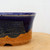 A detailed close-up of a Sean Guo American handmade bonsai pot, showcasing a striking two-toned glaze design. The top half of the ceramic pot is finished in a deep, glossy cobalt blue that artistically drips down over a textured, wave-like horizon line. Below this, the pot reveals its natural, warm orange-brown clay body, which is accented with dark mineral speckles for a rustic feel. The pot features a slightly flared rim at the top and a small, arched drainage hole visible at the very base. It is resting on a light-toned wooden surface against a clean, neutral white background, highlighting the artisanal craftsmanship and vibrant colors of the piece.