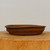 A shallow, oval-shaped unglazed bonsai pot handcrafted by American artist Paul Olson sits centered on a light-colored, natural wood pedestal. The pot is made from a rustic, earthy terracotta clay with a rich, reddish-brown color and subtle darker, scorched-looking accents around the flared rim. Its design features a low profile with a slight horizontal ridge running around the middle of its textured exterior, highlighting its artisanal quality. The pot is photographed from a side-view against a clean, off-white background, emphasizing the natural tones and handcrafted details of the ceramic piece.