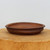A shallow, oval-shaped bonsai pot handmade from reddish-brown clay by American ceramicist Paul Olson. The pot has a rustic, unglazed finish with natural variations in texture and color, including dark speckles throughout. Its design features a pronounced, slightly rounded rim and a recessed horizontal band around the middle of the exterior wall. The pot is centered on a thick, cross-sectioned wooden slab with a natural grain, all set against a clean, off-white background that emphasizes its handcrafted details and earthy aesthetic.