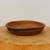A circular, shallow bonsai pot handcrafted from reddish-brown clay by artist Paul Olson. The pot features a wide, flat rim with a darker, scorched appearance and subtle horizontal ribbing along its exterior, characteristic of being hand-thrown on a potter's wheel. A single drainage hole is visible in the center of its smooth interior base. The pot sits on a thick, natural wooden block with a visible grain, set against a plain, light-colored background that emphasizes its rustic, artisanal quality. This American-made ceramic vessel exemplifies functional art for bonsai enthusiasts.