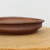 A detailed, side-profile close-up of an American handmade bonsai pot by artisan Paul Olson. This shallow, low-profile ceramic tray is crafted from unglazed, reddish-brown clay with a rustic, matte texture and visible dark speckles throughout the material. The exterior wall of the pot features horizontal throwing lines and a distinctive tiered or stepped design, leading up to a smooth, rounded rim. The pot sits on a light-colored, natural wooden surface, likely a display board or workbench, against a clean, out-of-focus white background that highlights the earthy tones and artisanal craftsmanship of the piece.