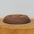 An upside-down view of a shallow, oval-shaped ceramic bonsai pot handcrafted by American artist Paul Olson. The pot is made from an earthy, reddish-brown clay with a matte, textured finish and fine dark specks throughout. Three large, clean-cut circular drainage holes are visible on the bottom, alongside a hand-carved artist's chop or signature in the center. The pot features a low profile with sides that curve inward toward the bottom and a thin, slightly flared rim at its base. It sits on a thick, light-colored wooden block with a visible natural grain, set against a plain, neutral background to emphasize the artisanal details of the piece.