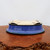 An oval-shaped glazed bonsai pot, shown upside down to highlight its design and functional features. The pot is finished in a vibrant, deep cobalt blue glaze with a high-gloss sheen that catches the light. The blue glaze extends down to a dark, almost black, slightly flared rim at what would be the base of the pot. Its unglazed, light tan ceramic bottom is visible, featuring four small integrated feet and drainage holes essential for maintaining the health of a bonsai tree by preventing waterlogging. The pot is resting on a polished, warm reddish-brown wood display stand with a natural, irregular edge. The entire composition is set against a minimalist, light grey background, focusing attention on the craftsmanship and rich color of this professional-grade ceramic bonsai container from Bonsai Outlet.