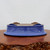 An inverted cobalt blue glazed ceramic bonsai pot rests on a dark, rustic wooden surface against a plain, off-white background. The pot, shown upside down to highlight its construction, features a vibrant and glossy cobalt blue glaze on its flared sides. The unglazed bottom is a natural terracotta-colored clay, revealing four small, integrated feet and multiple pre-drilled drainage and wire holes. Its smooth, oval shape and rich color make it a striking container for a variety of bonsai tree styles. The low-angle photograph emphasizes the pot's clean lines and high-quality finish, typical of professional bonsai pottery.