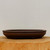 An oval-shaped, unglazed Yixing bonsai pot in a deep, earthy brown color, presented in a professional studio setting. The shallow pot features a smooth, matte finish with a subtle, rounded rim and sits on a recessed, footed base. It is placed centrally on a thick, light-colored natural wood slice, showcasing its rustic elegance against a clean, neutral off-white background. The simple yet sophisticated design of this Yixing clay pot highlights its traditional craftsmanship, making it an ideal choice for a variety of bonsai tree styles.