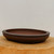 An elegant, high-quality Yixing unglazed bonsai pot (model 2451) featuring a deep, dark brown matte finish. The pot is shaped in a classic shallow oval with a wide, rounded rim that slopes slightly inward. It rests on small, integrated feet designed to provide essential drainage and air circulation for a bonsai tree. The pot is displayed on a rustic, light-toned wooden pedestal, creating a striking contrast that emphasizes its smooth texture and traditional craftsmanship. The background is a clean, neutral off-white, making this professional-grade ceramic piece the central focus.