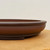 A close-up side view of a shallow, unglazed Yixing bonsai pot, model 2451, showcasing its fine craftsmanship and natural materials. The pot is crafted from a rich, reddish-brown "purple clay," featuring a smooth yet subtly grainy matte finish characteristic of traditional Yixing pottery. Its design includes a thick, rounded rim that curves elegantly outward, with a delicate horizontal groove running just beneath it for added visual interest. The body of the pot follows a gentle, convex curve, tapering down to a small, slightly inset foot at the base. The pot is positioned on a light-colored wooden surface against a soft-focus, off-white background, with even lighting that highlights the earthy tones and fine texture of the clay. This low-profile, unglazed ceramic pot is a classic choice for bonsai enthusiasts, offering both a beautiful aesthetic and a breathable environment for a tree's root system.