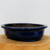 A deep cobalt blue glazed Yixing bonsai pot with a classic oval shape and a shallow profile, shown from a side-angle view. The pot features a wide, flat rim that flares out slightly and rests on small, inconspicuous feet. Its glossy glaze has a rich, multi-toned blue finish with subtle reflections. The interior of the ceramic pot is a dark, unglazed earthy brown. It is placed on a light-colored, natural-edged wooden base against a minimalist off-white background, emphasizing its elegant form and craftsmanship.