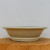 A side-profile view of a round Yixing glazed bonsai pot with a light beige or cream-colored finish. The pot features a wide, flared rim that tapers down to a sturdy, tiered base, creating an elegant and classic silhouette. Its surface has a smooth, glossy glaze with subtle speckles and tonal variations characteristic of authentic Yixing ceramic craftsmanship. The pot is displayed on a thick, natural wood block with visible grain, set against a plain, neutral-toned wall to highlight its form and neutral color palette. This type of high-quality bonsai container is designed to complement a variety of tree species and styles.