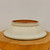 A high-resolution product photograph captures a shallow, round Yixing glazed bonsai pot, shown upside down to highlight its shape and construction. Resting on a thick, rustic wooden block with a natural grain, the pot features a smooth, pale off-white or light celadon glaze accented by subtle, tiny green speckles. 

The pot’s design is characterized by a wide, elegantly flared rim that tapers inward towards a narrower base. A fine, raised ridge encircles the body just above the flared lip, adding a classic architectural detail. The unglazed base, visible at the top of the image, reveals the natural, warm reddish-brown clay synonymous with traditional Yixing pottery. A large, circular drainage hole is clearly visible in the center of the base, essential for healthy bonsai root systems. The composition is set against a clean, neutral light-gray background, focusing all attention on the craftsmanship and delicate color of this ceramic bonsai planter.