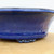 A detailed close-up shot of a deep cobalt blue glazed Yixing bonsai pot, highlighting its rich color and professional finish. The image focuses on the side profile of the ceramic pot, showing a smooth, lustrous glaze with subtle variations in tone. Near the base, a single decorative "cloud" foot is visible, featuring intricate scrolling details that are also covered in the same glossy blue glaze. The pot has a slight outward flare at the top rim and a subtle horizontal ridge near the bottom edge. It sits on a light-colored tan wooden surface against a clean, neutral white background, emphasizing the elegant craftsmanship of this Yixing pottery.