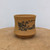 A detailed view of a light-brown, unglazed Yixing bonsai pot, showcasing its sandy, matte texture and elegant shape. The ceramic container features a slightly waisted middle that flares out towards a footed base. The front of the pot is decorated with a traditional Chinese landscape illustration in black ink, depicting ancient pine trees, a hillside building, and a flowing river against a mountainous backdrop. Above the landscape art is a line of classic Chinese calligraphy. The pot is positioned on a rustic, light-toned wooden surface against a plain, off-white background, highlighting the natural beauty of the Zisha clay.