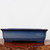A side-view studio photograph of a rectangular glazed ceramic bonsai pot in a stunning deep cobalt blue finish. The pot’s glaze exhibits a beautiful gradient, with richer, darker tones at the flared rim that softly fade into a lighter, dusty blue with fine mottling near the base. It features sturdy, integrated corner feet that provide essential height for drainage. The glossy pot sits atop a polished reddish-brown wood stand with a natural, irregular burl edge, showcasing the container's craftsmanship against a minimalist, neutral white background.