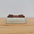 An upside-down view of a rectangular Yixing bonsai pot (model no. 2440) showcasing its construction and finish. The pot features a smooth, light cream or pale celadon-colored glaze on its exterior walls, which contrasts with the unglazed, dark reddish-brown Yixing clay on its base. The pot is designed with four decorative feet at the corners and two large drainage holes for optimal soil aeration and water management. It has rounded edges and slightly inward-tapering sides, resting on a light-colored wooden tabletop against a plain, off-white wall.