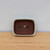 A top-down, high-angle view of a rectangular Yixing bonsai pot with rounded corners. The pot features a thick, cream-colored glazed rim that contrasts with its unglazed, deep reddish-brown clay interior, a hallmark of Yixing pottery. Inside the base of the pot, two large circular drainage holes are centrally positioned, accompanied by four smaller wire holes near the corners for securing a tree. The pot is centered on a light-colored wooden surface with a visible grain, set against a plain, neutral-toned wall.