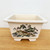 A high-angle, front-facing shot shows a 9-inch glazed Yixing bonsai pot, ideal for cascade or semi-cascade bonsai styles. The pot is a creamy off-white color with a square shape, a wide flared rim, and four small rectangular feet at the base. It sits on a light-grained wooden tabletop against a plain white background. 

The front side of the pot features an intricate, hand-painted traditional Chinese landscape scene. The painting depicts mist-covered, rugged mountains in the background with a small village of thatched-roof houses nestled in the valley. In the foreground, a large, gnarled pine tree stands next to smaller trees with pink blossoms, reminiscent of cherry or plum blossoms. The scene is rendered in a "Shan Shui" style with a palette of earthy greens, blues, browns, and subtle pops of red and pink, capturing a peaceful, ancient wilderness aesthetic.