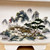 A close-up view of the intricate hand-painted artwork on a white, glazed Yixing bonsai pot. The traditional Chinese landscape painting, done in a classic shanshui style, depicts a serene mountain scene. In the background, towering, craggy peaks in shades of blue and grey are partially shrouded in soft, white mist. 

In the mid-ground, a cluster of small houses with thatched roofs sits on a rocky outcrop. Two small figures dressed in traditional red and white robes stand outside the buildings. To the right of the houses, a large, gnarled pine tree with dense green needles and a weathered trunk leans over the scene, accompanied by smaller trees bursting with delicate pink blossoms, suggestive of cherry or plum trees in spring. 

In the lower-left foreground, a solitary figure in a small, simple boat poles across a calm body of water. The rocky shoreline is dotted with patches of green moss and small shrubs. The painting uses a muted color palette with pops of vibrant red and pink, all set against the smooth, off-white surface of the rectangular ceramic pot, which features two small, blocky feet at the base.